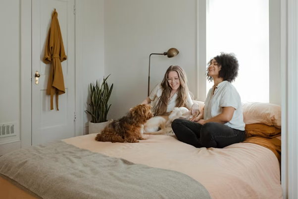 Two women sit on a bed with a small dog.